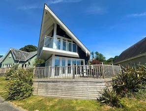 Ein modernes, zweistöckiges Haus mit Balkon, umgeben von einer Holzterrasse und viel Grün.