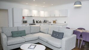 A modern living room with a grey sectional sofa, a white coffee table, and a kitchen area with white cabinets and appliances. - Seaforth (Isle of Lewis)