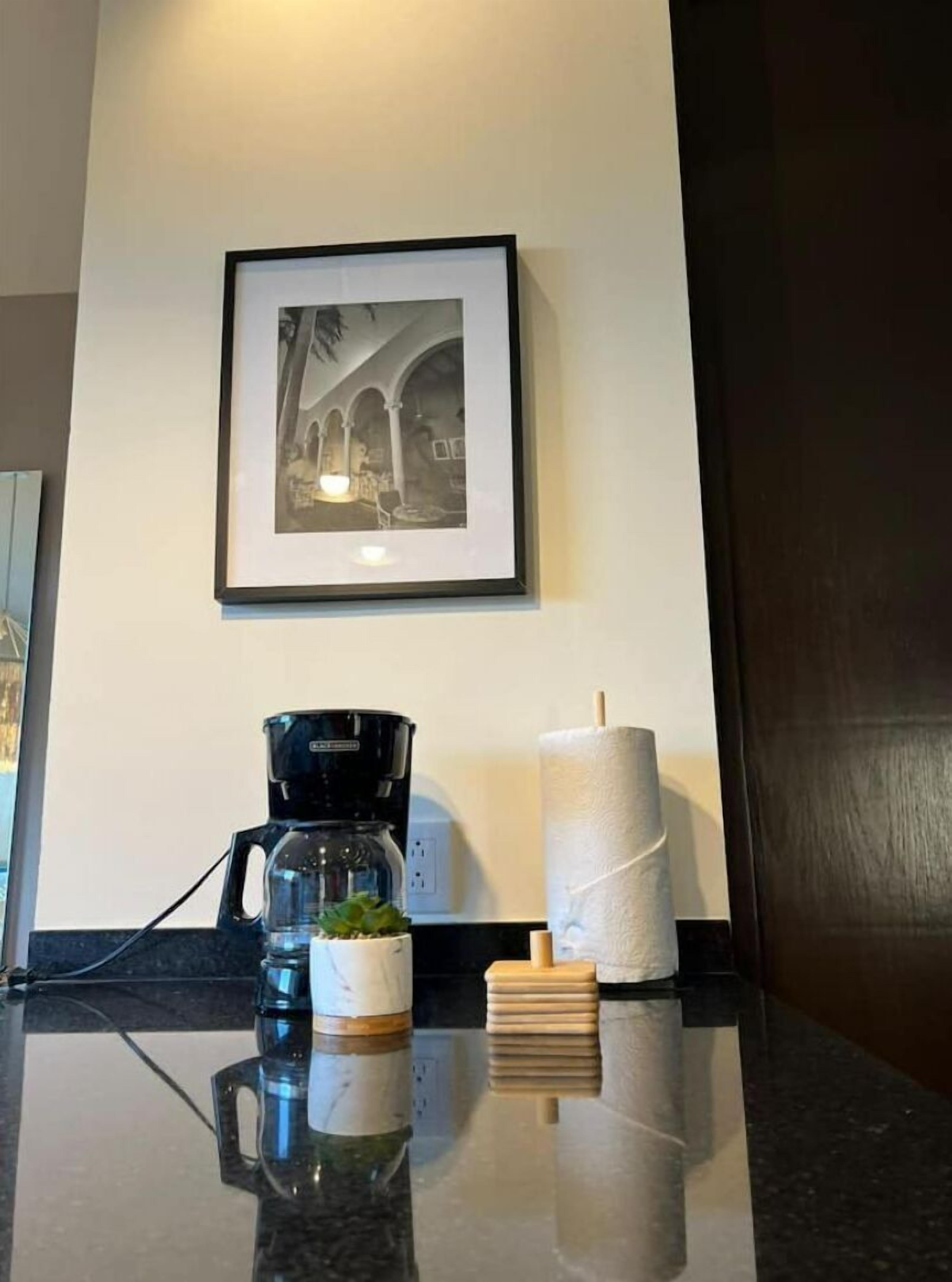A framed black and white photograph on the wall, a coffee maker, a roll of toilet paper, and a small plant on a reflective surface.