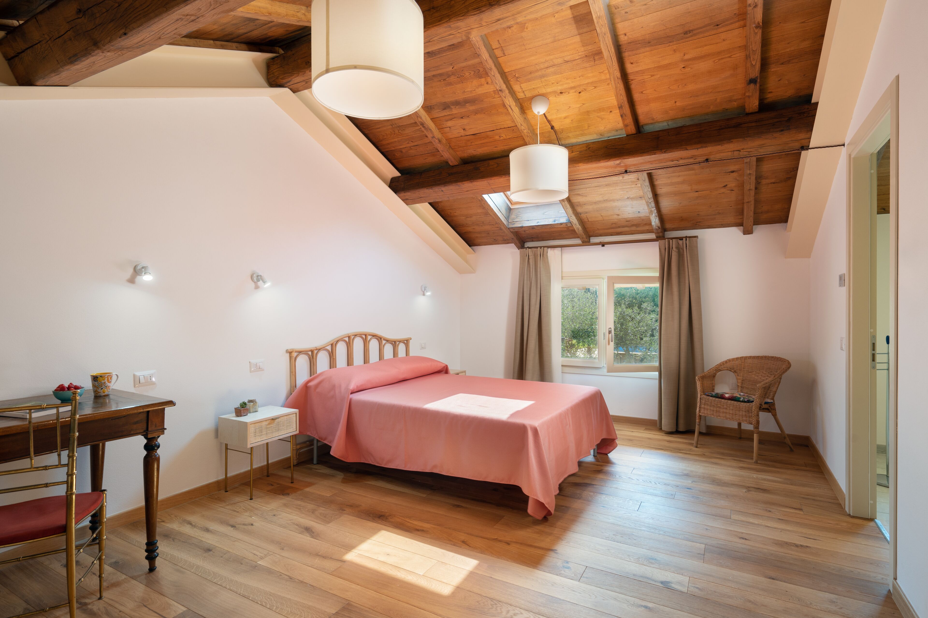 Une chambre avec un plafond en bois, un lit recouvert d’une couverture rose, un bureau en bois avec une chaise et une fenêtre avec des rideaux.