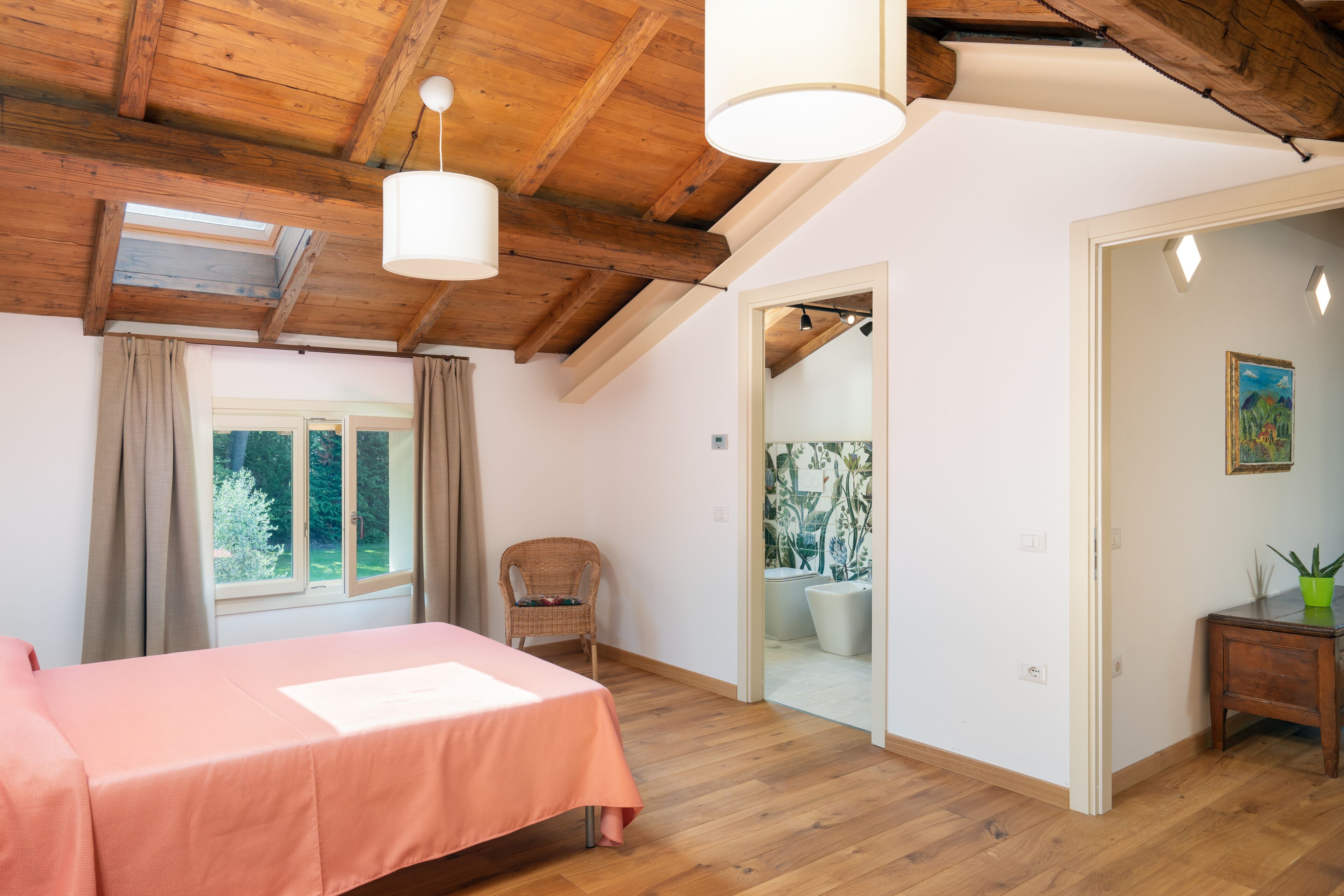 A bedroom with a wooden ceiling, a bed with a pink bedspread, a wicker chair, and a small table with a plant.