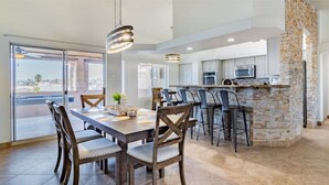 Un comedor amplio con una mesa grande y sillas, una cocina con un muro decorativo de piedra y vistas a través del ventanal.