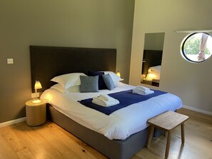 Ein ordentlich bezogenes Bett mit weißen Laken und einer blauen Decke, eine Nachttischlampe und ein runder Spiegel an der Wand.