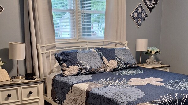 Une chambre à coucher comprenant un lit, des tables de chevet, une fenêtre avec des rideaux, un ventilateur de plafond et des œuvres d’art décoratives au mur.