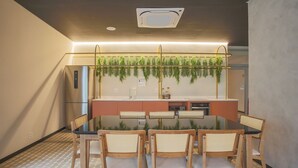 Een moderne eethoek met een glazen tafel en houten stoelen, een keukentoog met planten en een plafondlamp.