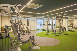 Un gimnasio con diversas máquinas de ejercicio, piso de césped artificial verde y un amplio ventanal que proporciona luz natural.