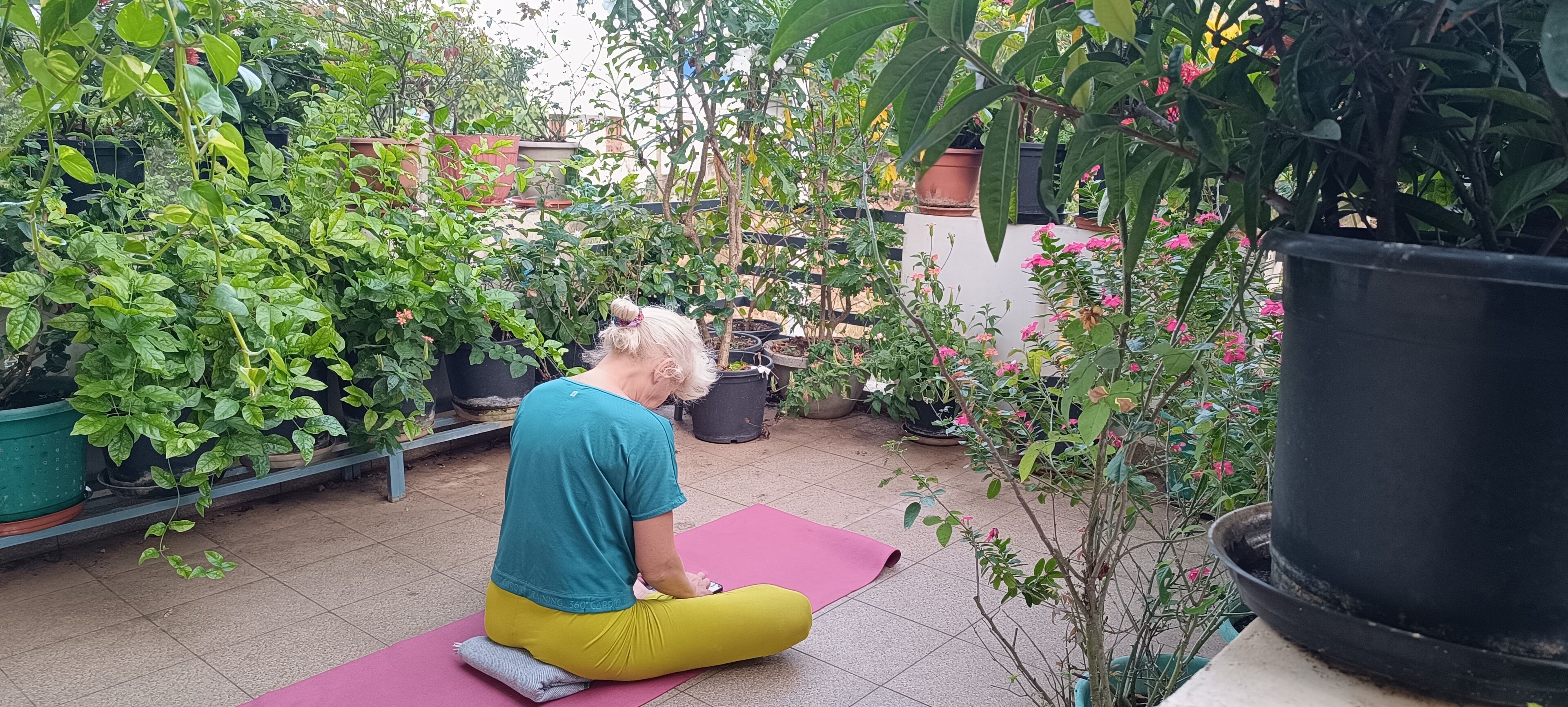 A person sitting on a yoga mat in an outdoor area with potted plants and a tiled floor.