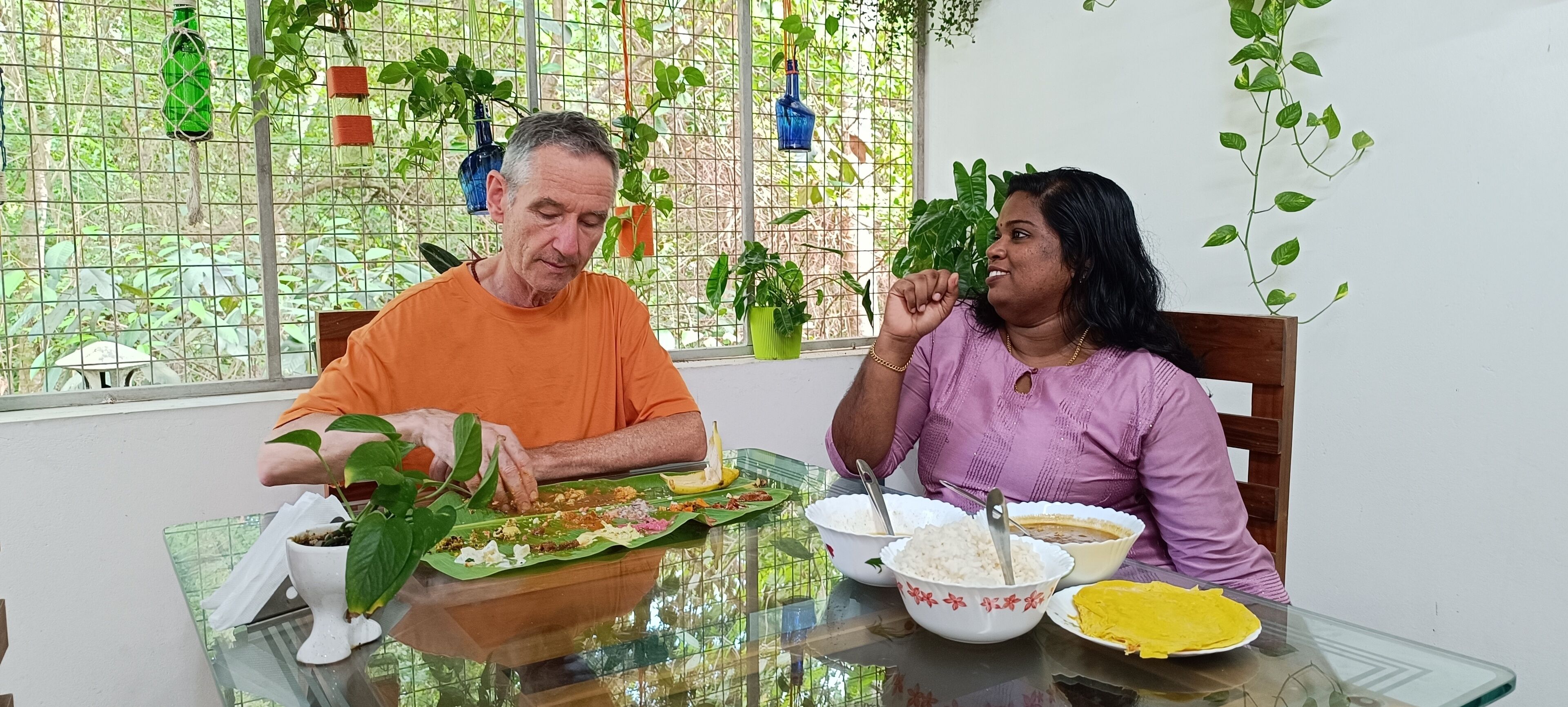 Dos personas cenando en una mesa con una variedad de platos, incluyendo arroz, curry y un plato amarillo, en una habitación con ventana y plantas en macetas.