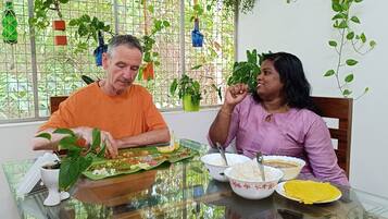Dos personas cenando en una mesa con una variedad de platos, incluyendo arroz, curry y un plato amarillo, en una habitación con ventana y plantas en macetas.