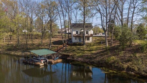 Una casa con un muelle que se adentra en un lago tranquilo, rodeada de árboles y un césped.