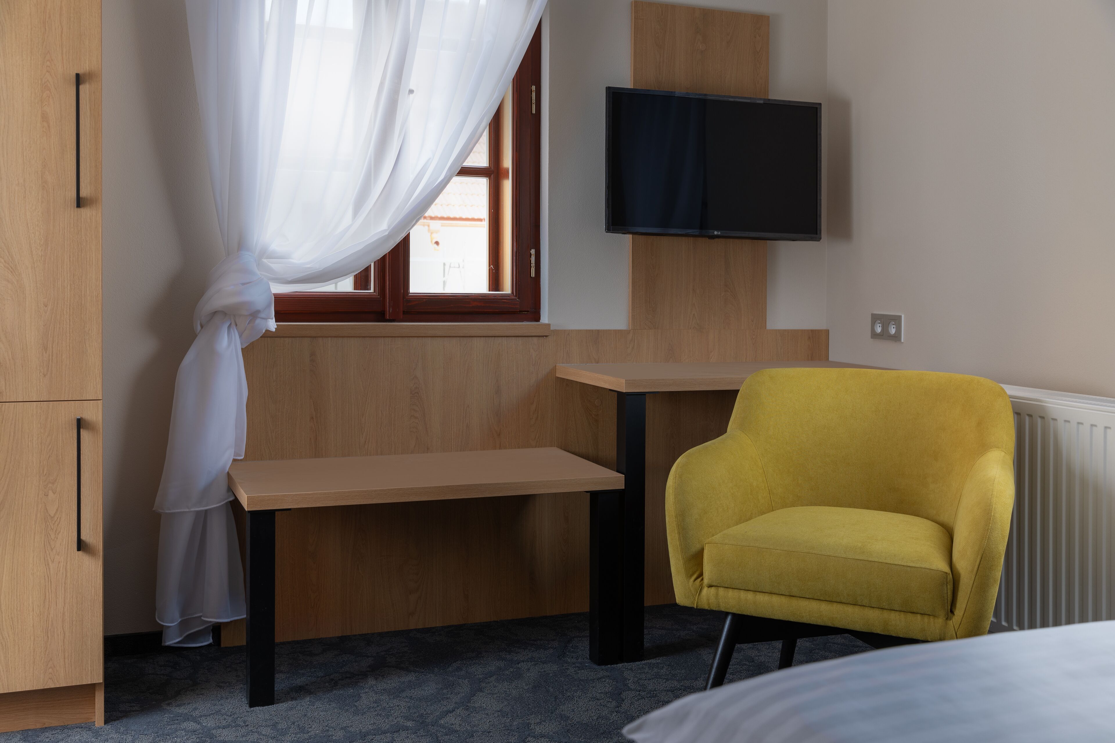 A room with a bed, a desk, a yellow armchair, a TV mounted on the wall, and a window with curtains.