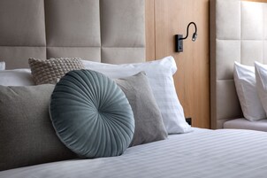 Una cama bien tendida con cabecera, una lámpara fijada a la pared y un panel de madera detrás de la cabecera.