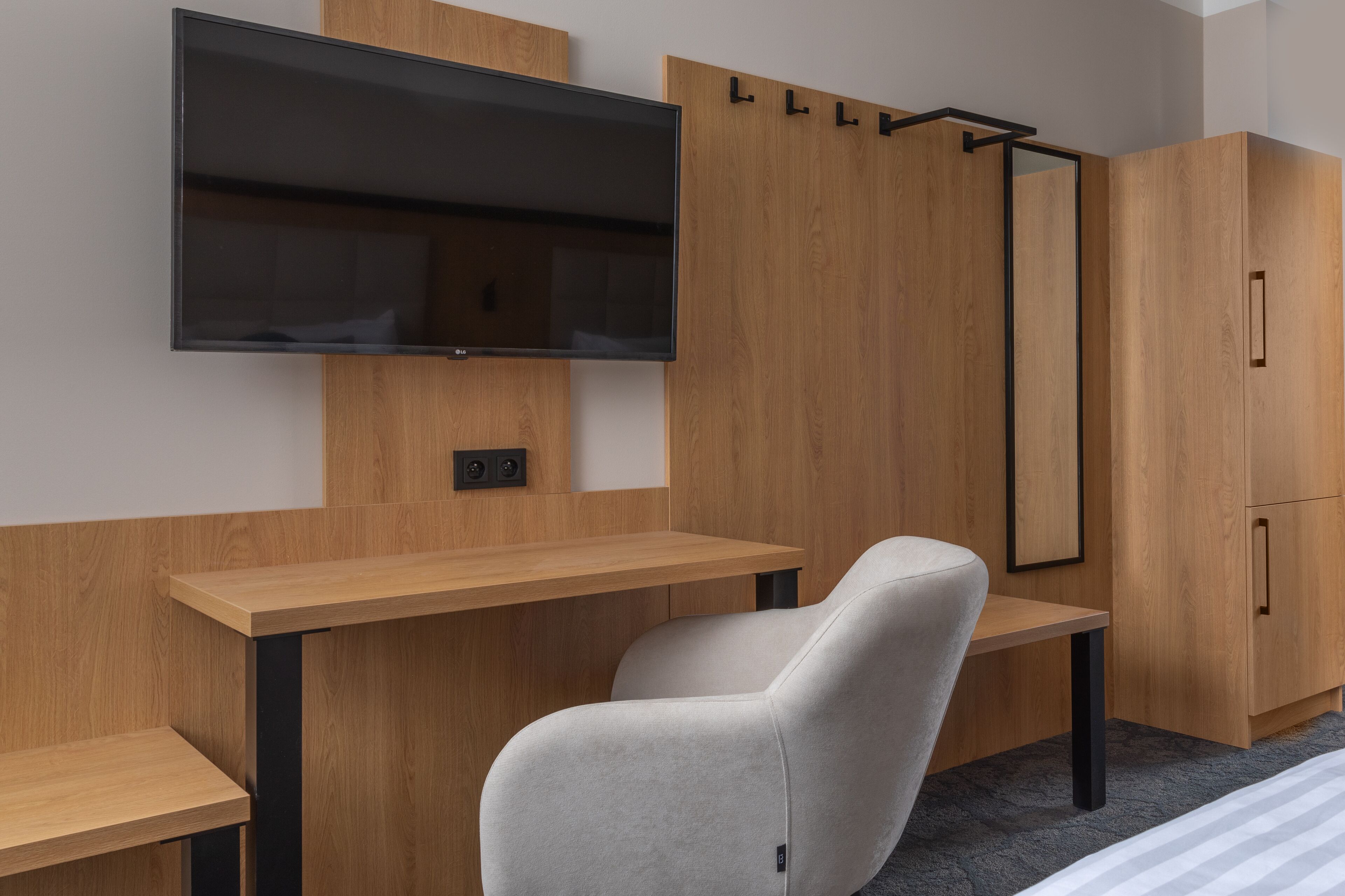 Una habitación moderna con un televisor de pantalla plana montado en la pared, un escritorio de madera, una silla gris y un armario con espejo.