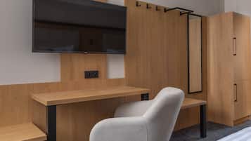 A modern room with a flat-screen TV mounted on the wall, a wooden desk, a grey chair, and a wardrobe with a mirror.