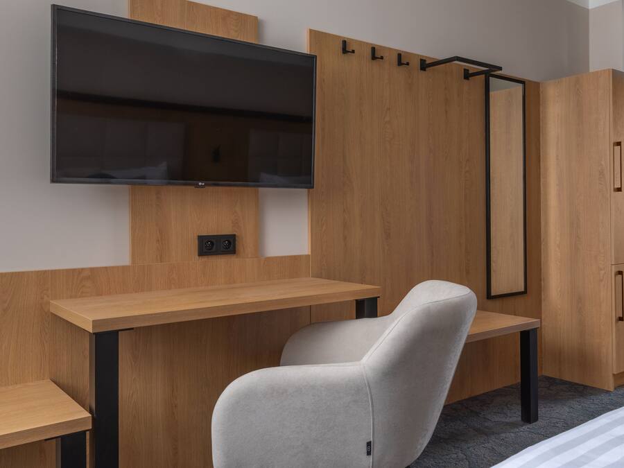 A modern room with a flat-screen TV mounted on the wall, a wooden desk, a grey chair, and a wardrobe with a mirror.