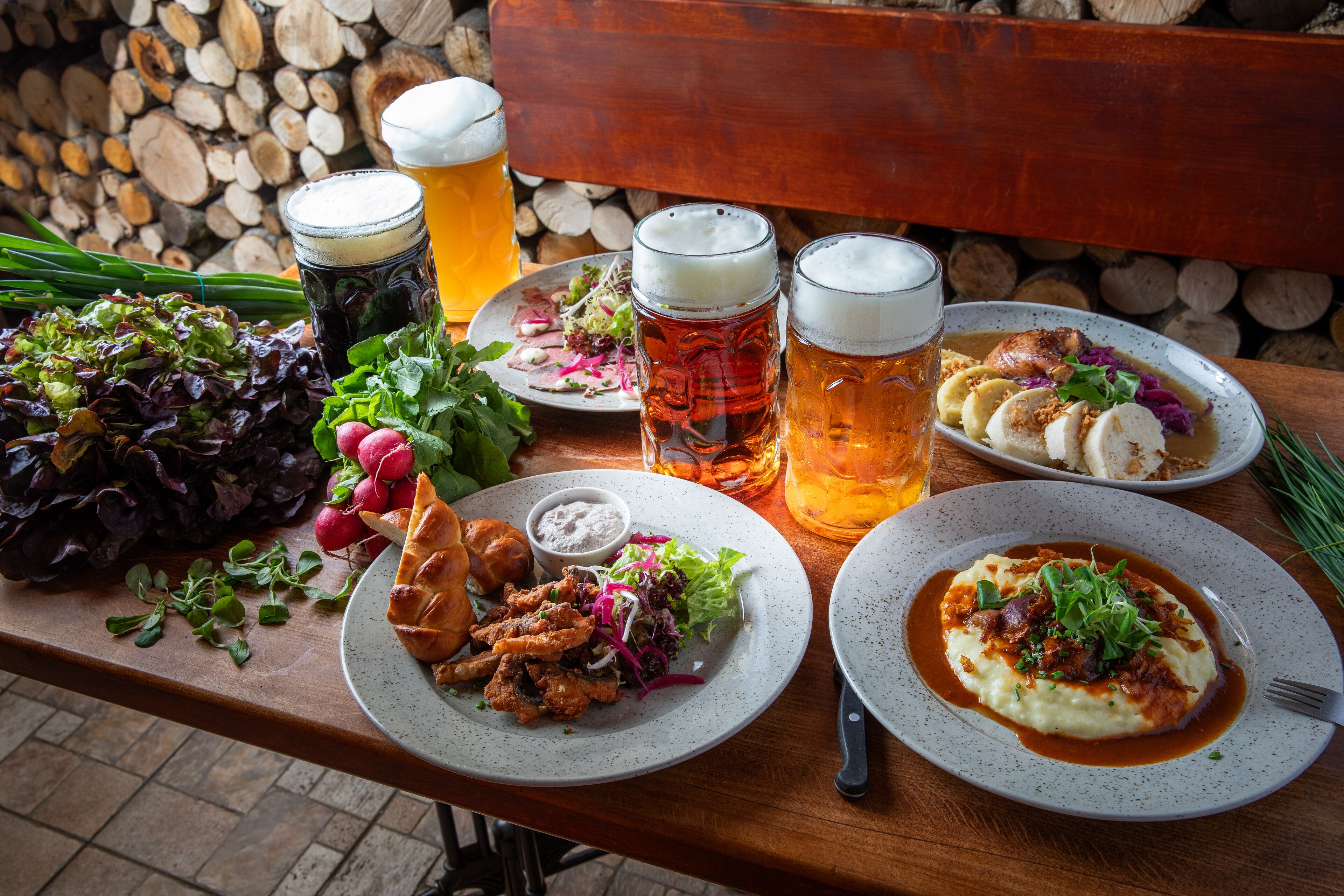 A wooden table with plates of food, beer mugs, and a salad.