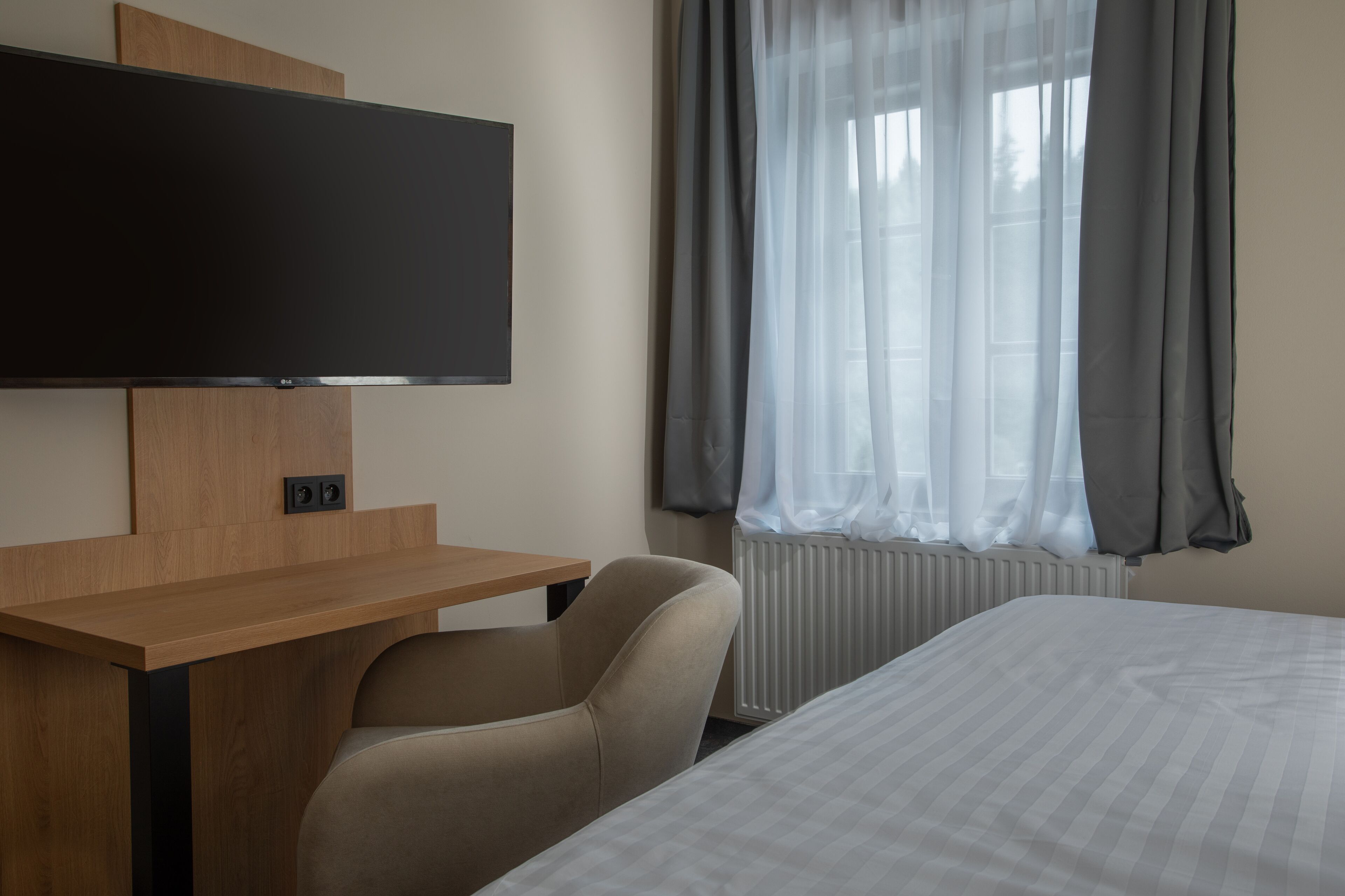 A hotel room with a bed, a desk with a flat-screen TV, a chair, and a window with curtains.