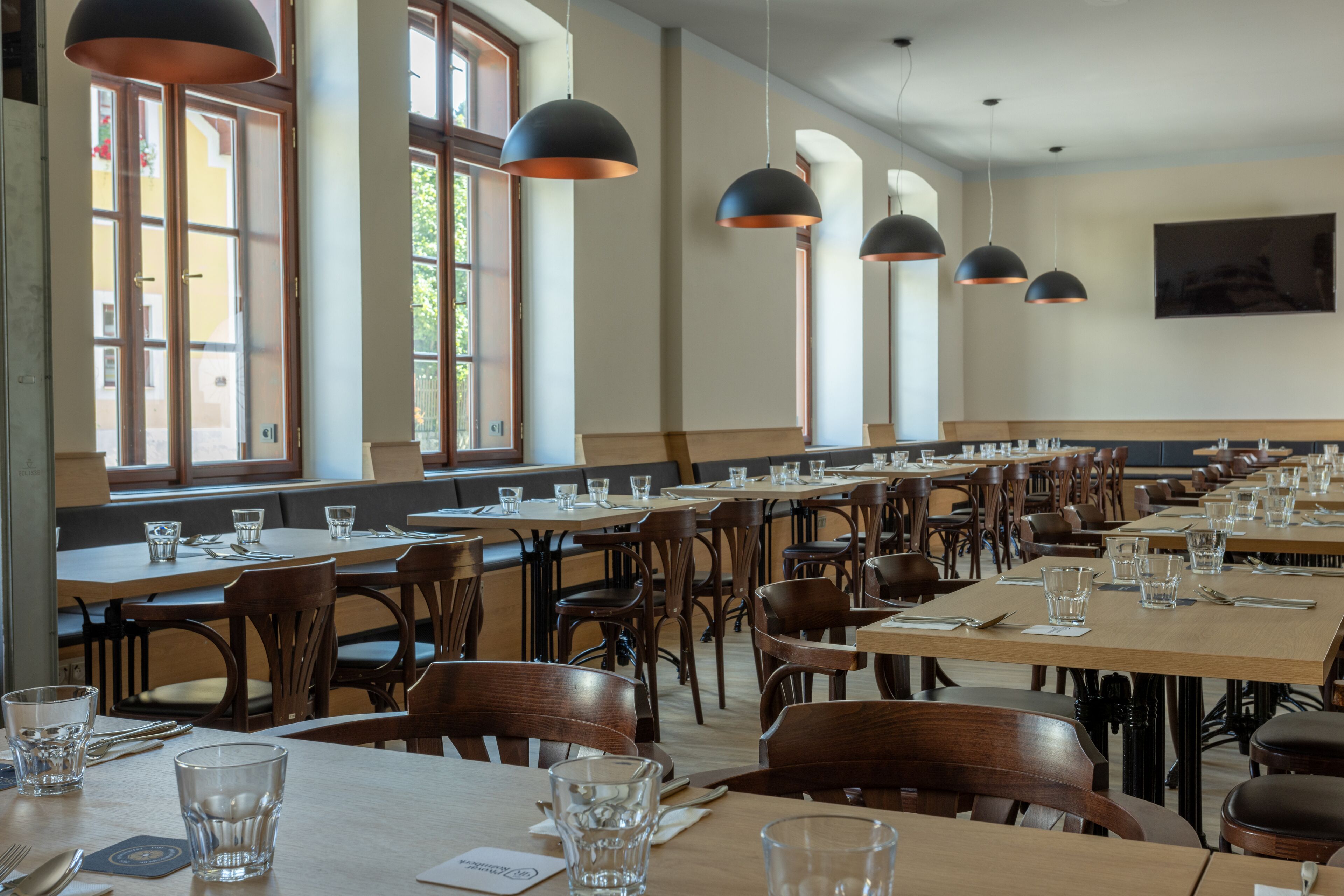 A spacious dining area with multiple tables and chairs, large windows, and pendant lighting.