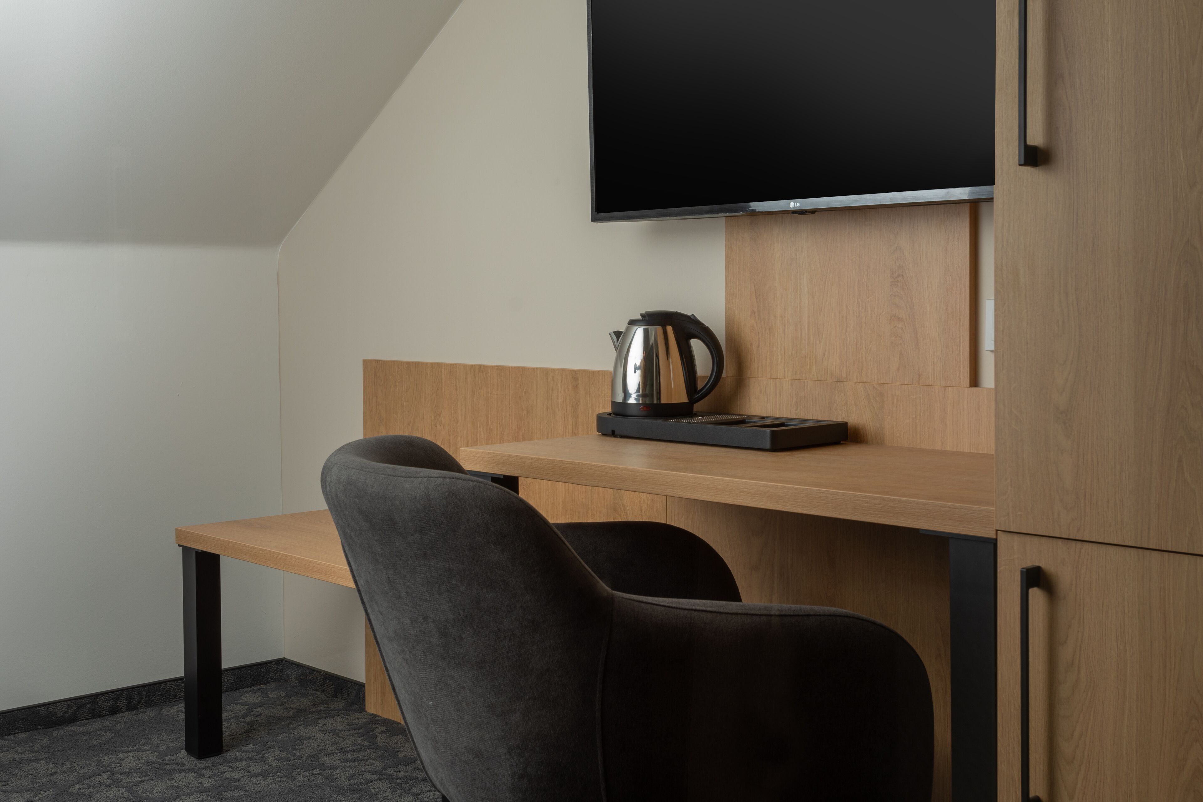 Une chambre moderne avec un bureau en bois, deux chaises noires, un téléviseur à écran plat fixé au mur et une machine à café posée sur le bureau.