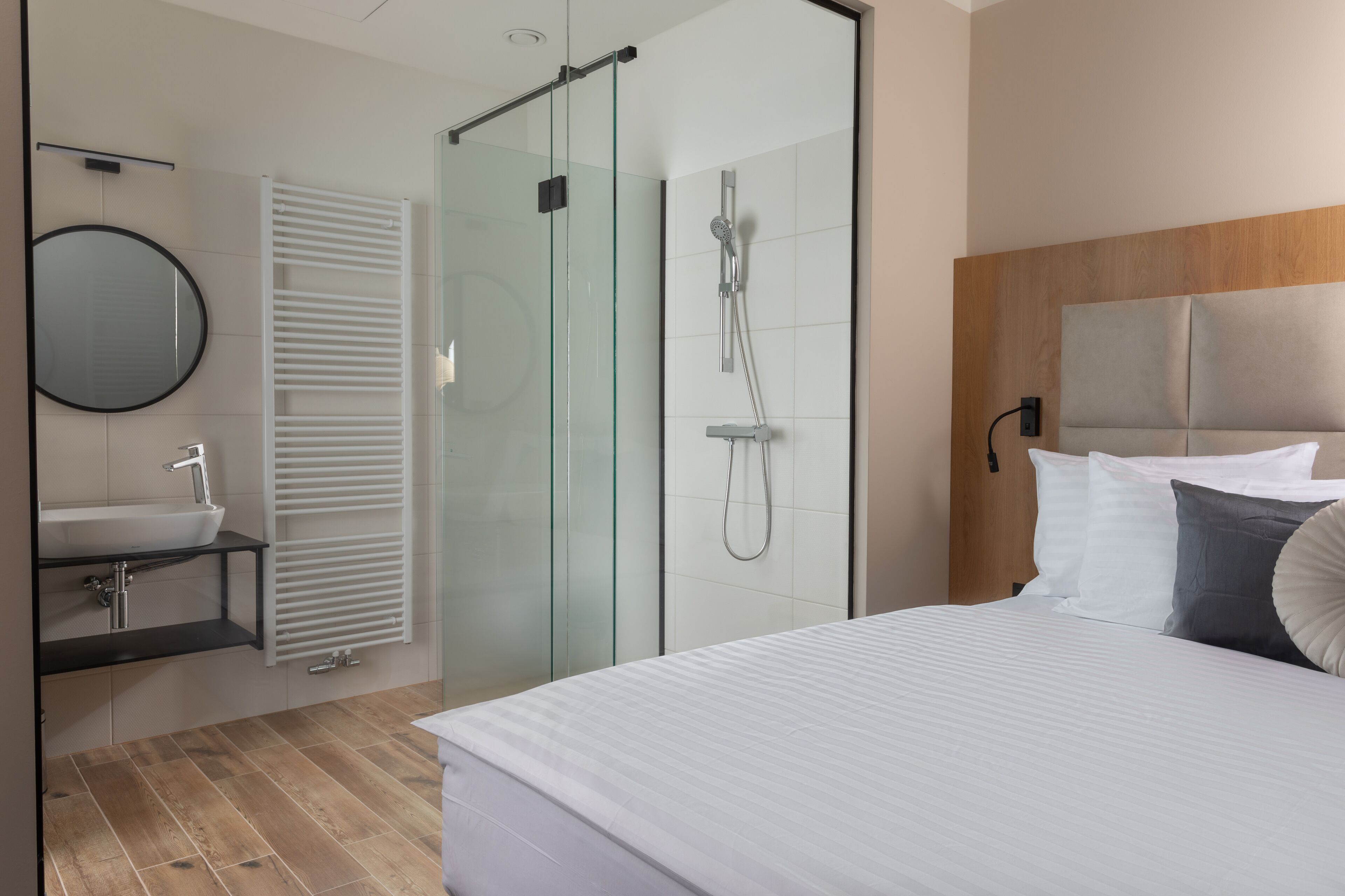 A modern hotel room with a glass-enclosed shower, a wooden headboard, a large bed, and a round mirror.