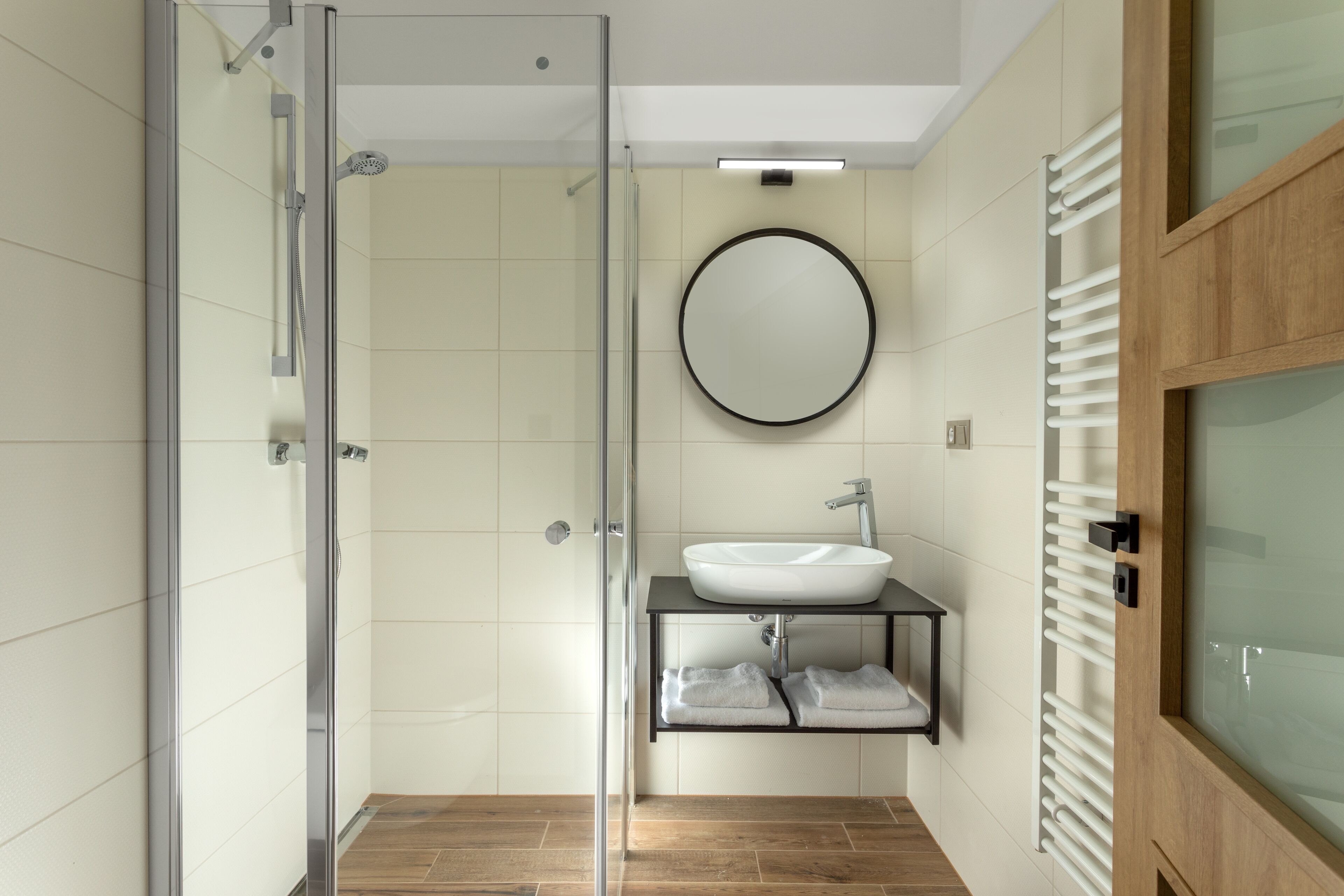 Een moderne badkamer met een glazen douchecabine, een ronde spiegel, een witte wastafel en een handdoekrek.