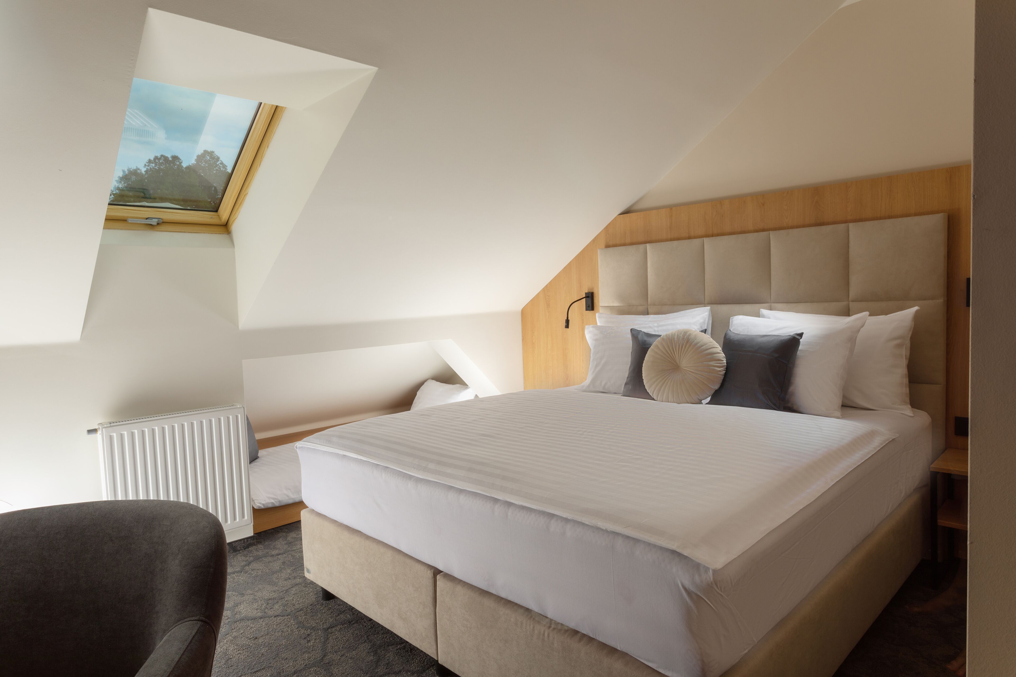 Quarto moderno com cama grande, cabeceira de madeira, claraboia e radiador.