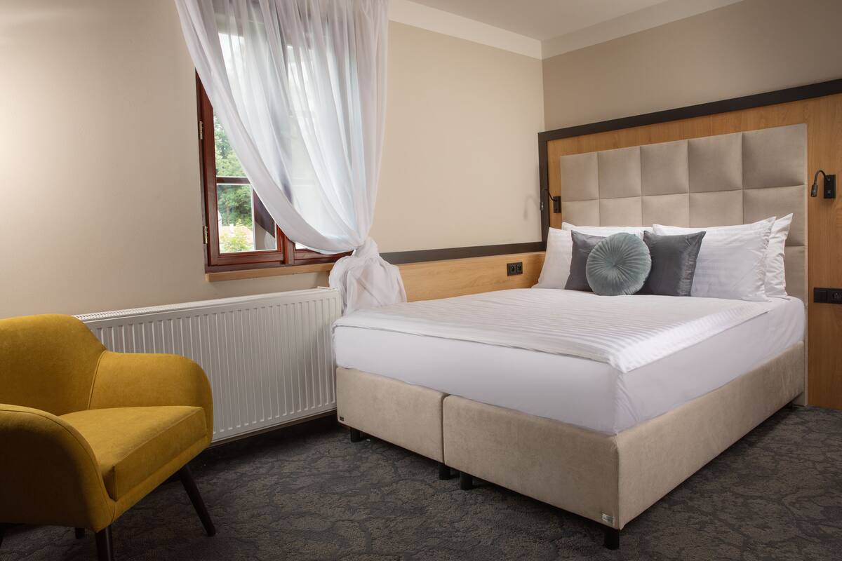 A hotel room with a bed, a chair, a window with curtains, and a radiator.