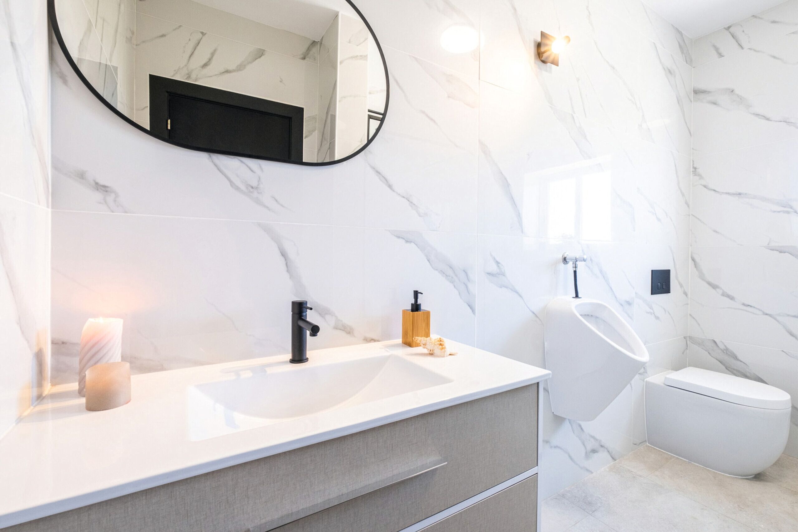 A bathroom with a white marble sink, a black faucet, a round mirror, a toilet, and a wall-mounted light fixture.