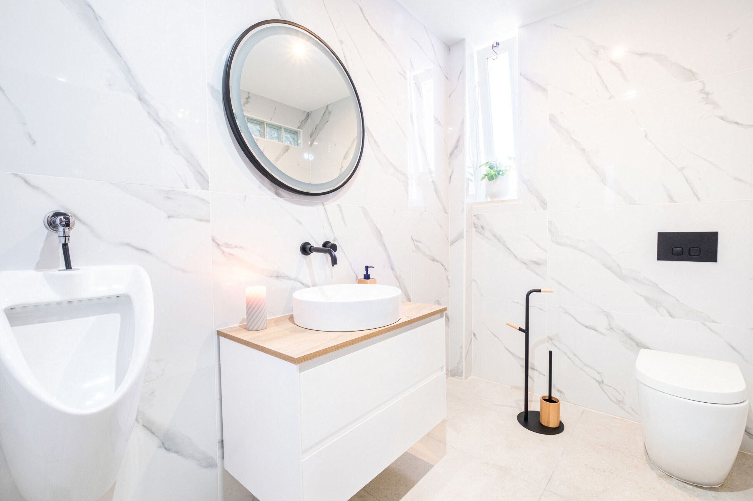 A modern bathroom with a marble wall, a round mirror, a white sink, a toilet, and a bidet.