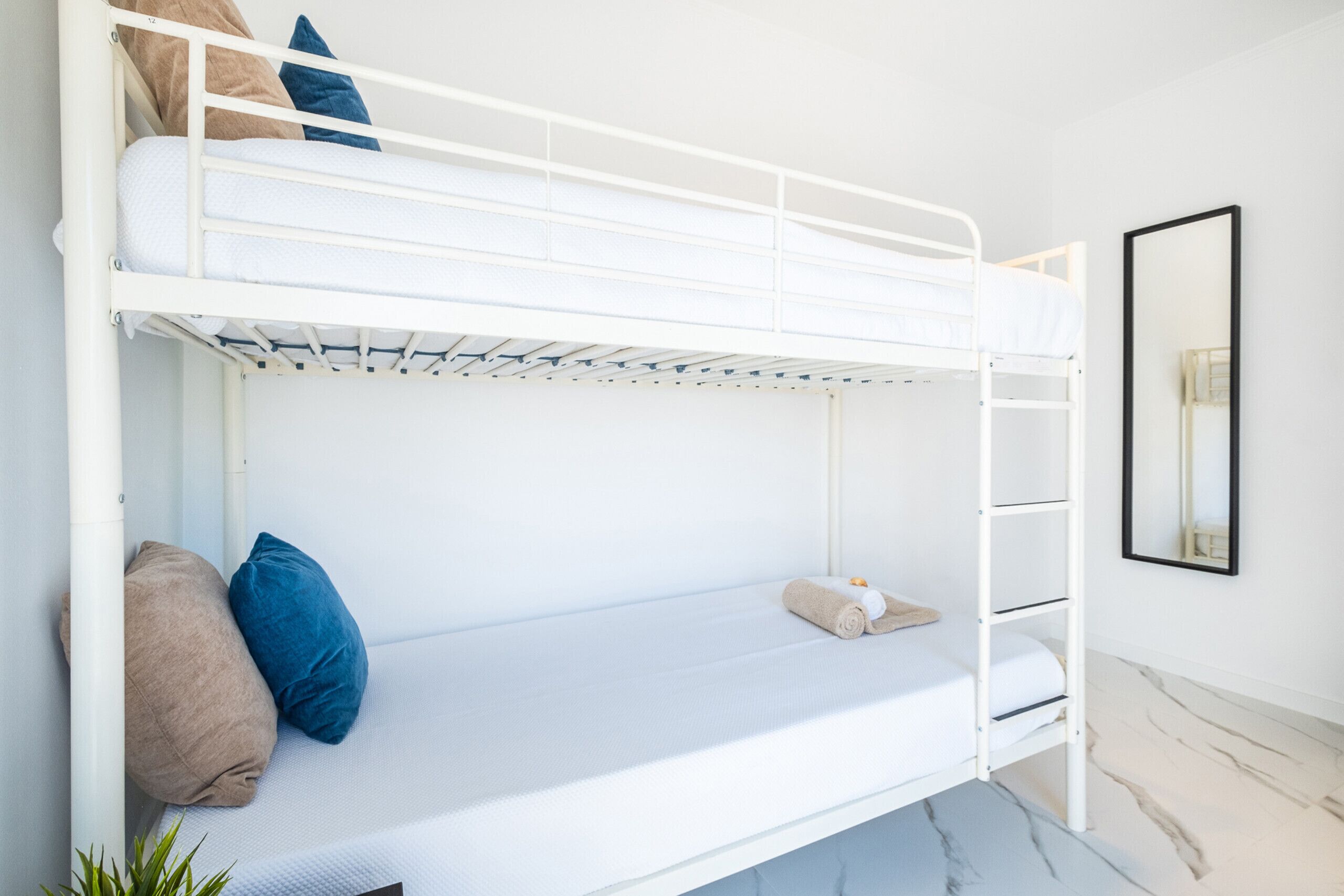 Una cama litera con estructura blanca, una plaza inferior con colchón y almohadas, y una plaza superior con colchón y almohadas.