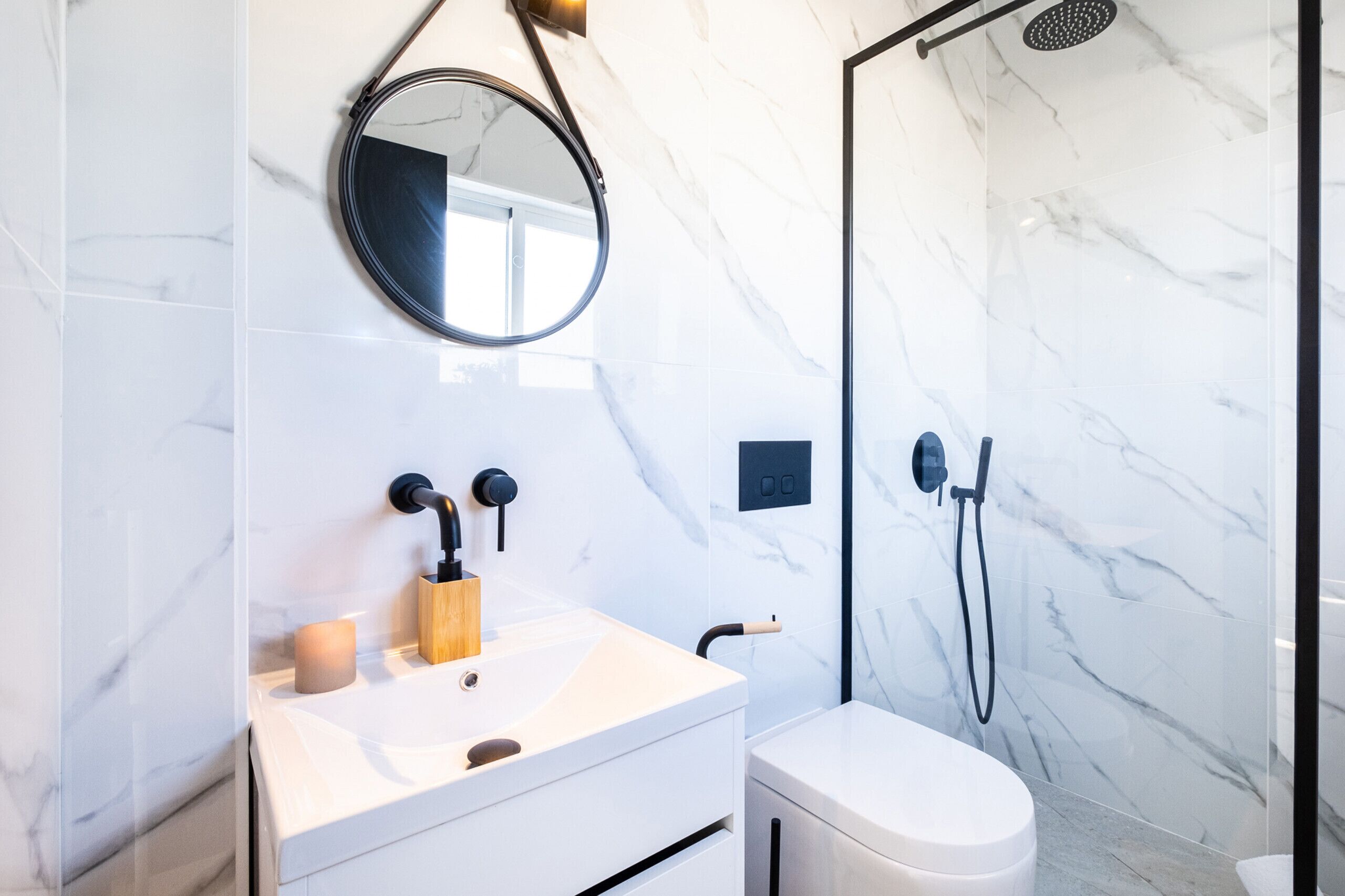 A modern bathroom with a marble wall, a round mirror, a sink with a wooden soap holder, and a glass-enclosed shower.
