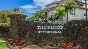 Een stenen muur met de aanduiding 'THE VILLAS AT POIPUKAI', een groot huis met pannendak en palmbomen.