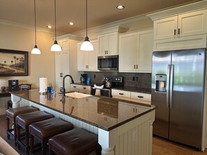 A modern kitchen with white cabinetry, a stainless steel refrigerator, a granite countertop, and three bar stools.