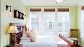 Habitación de hotel con cama, una silla, una mesita de noche, una lámpara y ventana con vista a un balcón.