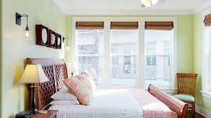 Habitación de hotel con cama, una silla, una mesita de noche, una lámpara y ventana con vista a un balcón.