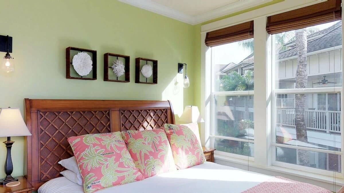 Une chambre à coucher avec une tête de lit en bois, un lit agrémenté d’oreillers à motifs floraux, une table de chevet avec une lampe et une fenêtre donnant sur un quartier résidentiel.