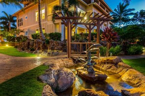 A well-maintained garden with a water feature, a stone pathway, and a building with a balcony in the background. - 3BR I Luxury in Paradise l Beaches, Golf, Shopping (Koloa)