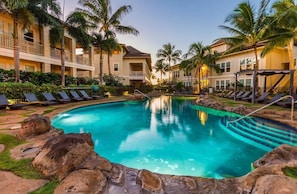 Uma área de piscina de hotel com espreguiçadeiras, palmeiras e uma piscina azul cristalina rodeada por pedras.