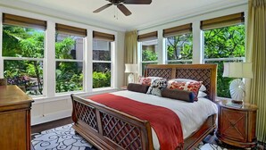 Un dormitorio con cama, ventilador de techo, ventanas amplias con persianas y vistas a zonas verdes.