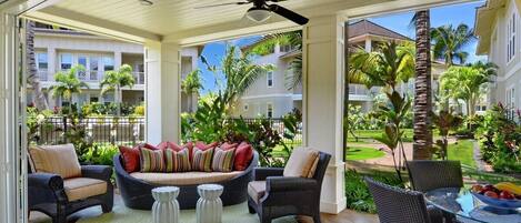 A covered outdoor seating area with wicker furniture, a ceiling fan, and a view of a lush garden and buildings in the background.