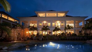 Un hotel grande de varios pisos con piscina, iluminación exterior y palmeras.