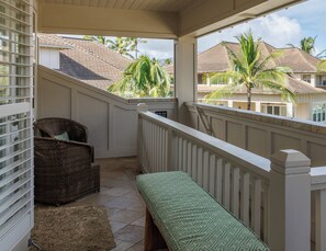 A balcony with a wicker chair, a bench, and a view of palm trees and houses.
