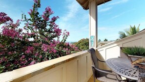 Varanda com mesa e cadeiras, vista para um jardim com flores cor-de-rosa e céu azul.