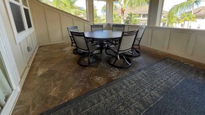 Une terrasse couverte avec une table ronde et des chaises, de grandes fenêtres et une vue sur les palmiers et les bâtiments.