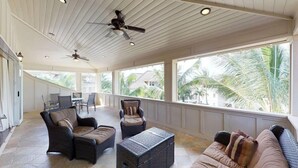 Un espace intérieur spacieux, doté d’un ventilateur de plafond, de meubles en osier et de grandes fenêtres offrant une vue sur les palmiers.