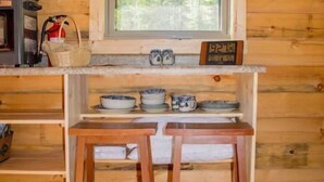 A wooden cabin interior with a kitchen area featuring a window, a shelf with dishes, and two wooden stools.