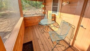A wooden cabin with a porch, two metal chairs, a small table, and a window overlooking a forested area.