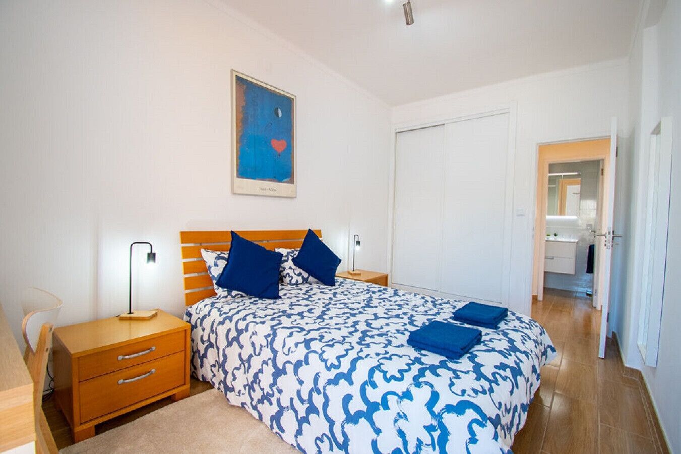 A bedroom with a bed, bedside tables, a nightstand, a wall-mounted lamp, and a door leading to a bathroom.