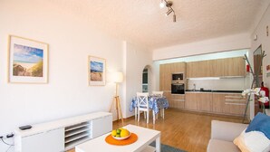 Una sala de estar moderna con un sofá blanco, una mesa de comedor con un tazón de fruta y una cocina con armarios de madera y un televisor.