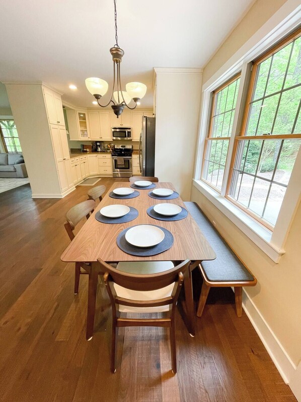 A dining area with a wooden table set for four, surrounded by chairs, with a bench along one side. A chandelier hangs from the ceiling, and a window provides a view of greenery outside.